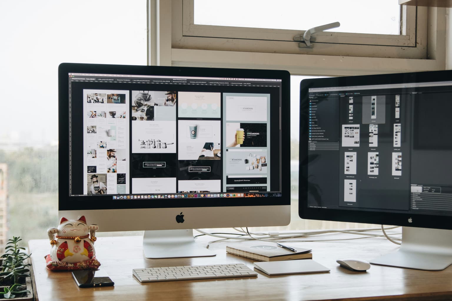 Two Apple desktop computers on a wooden desk display website design layouts in Photoshop, with a lucky cat ornament, keyboard and mouse nearby.