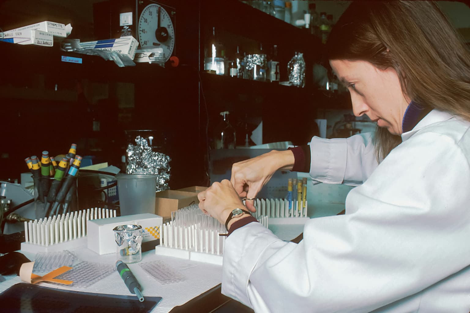 A woman in a white lab coat handles test tubes at a laboratory bench cluttered with pipettes, foil-wrapped items and a microplate.